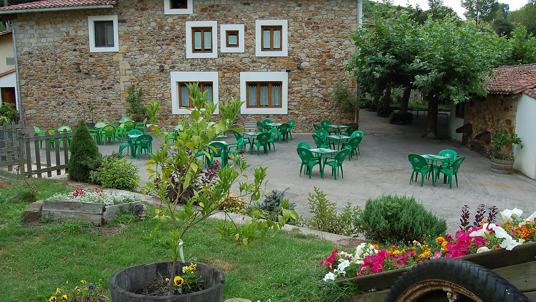 El caserío de Larrinagatxu visto desde el jardín, con las flores delante y las mesas verdes de la terraza al fondo.
