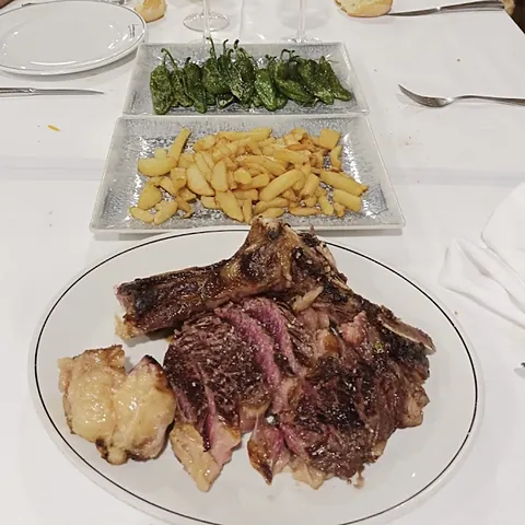 Chuletón cortado sobre el plato, con patatas y pimientos de padrón al lado.